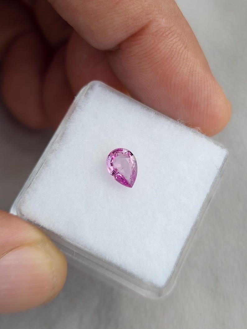 Side angle of the 0.75ct pink sapphire, unheated and natural. The pear cut loose gemstone glows with vivid magenta rose hues, showing well-balanced symmetry and brilliance. Ideal for bespoke jeNatural sapphire placed on a white jewelry box pad held in hand, pear cut vivid orchid pink color, unheated Madagascar sapphire shown for true size and setting scale, suitable for custom jewelry.welry design, pendants, or gemstone collections.