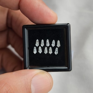 Pode incluir: Uma caixa de joias quadrada preta contém uma coleção de pequenas pedras preciosas claras e facetadas. As pedras preciosas estão dispostas em duas fileiras de cinco, cada uma com uma forma alongada única. A caixa é segurada contra um fundo branco macio.