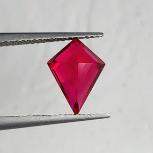 Front view of the same flawless lab created ruby in tweezers over a light backdrop. The kite shape silhouette is clean and symmetrical, with zero visible inclusions and vivid pigeon blood red color. This high-end man-made ruby is cut for luxury jewelry buyers who want dramatic color, modern geometry, and perfect clarity for bespoke pieces.