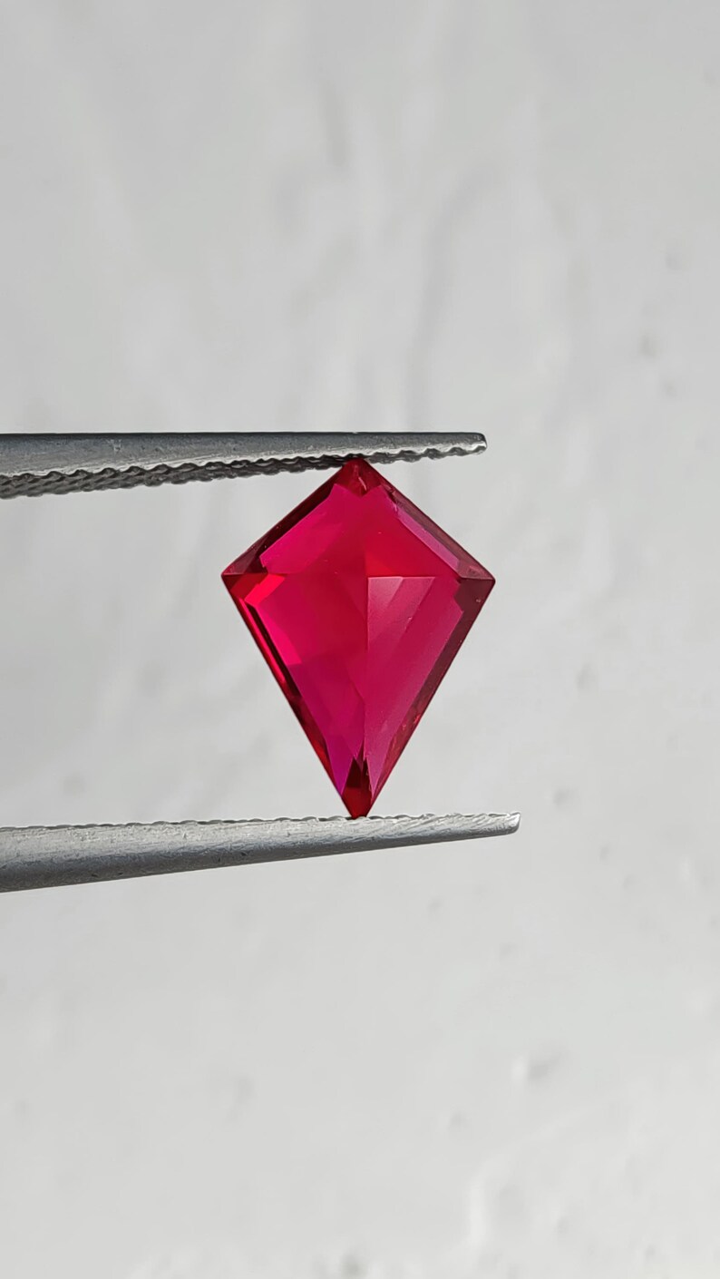 Front view of the same flawless lab created ruby in tweezers over a light backdrop. The kite shape silhouette is clean and symmetrical, with zero visible inclusions and vivid pigeon blood red color. This high-end man-made ruby is cut for luxury jewelry buyers who want dramatic color, modern geometry, and perfect clarity for bespoke pieces.