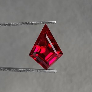 Loose lab grown ruby in a geometric kite cut held in gem tweezers against a neutral gray background. The stone shows vivid pigeon blood red color with clean step-style facets and razor meet points. Flawless clarity and high Thai polish make it ideal as a custom engagement ring center stone, pendant focal, or luxury earring pair. Precision-cut AAA grade synthetic corundum, ready for jewelry.