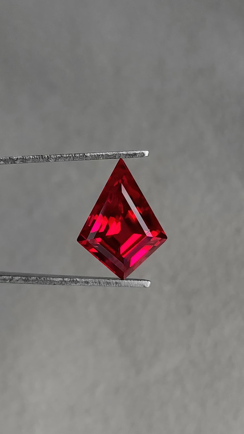 Loose lab grown ruby in a geometric kite cut held in gem tweezers against a neutral gray background. The stone shows vivid pigeon blood red color with clean step-style facets and razor meet points. Flawless clarity and high Thai polish make it ideal as a custom engagement ring center stone, pendant focal, or luxury earring pair. Precision-cut AAA grade synthetic corundum, ready for jewelry.
