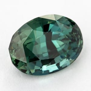 Puede incluir: Una piedra preciosa de corte ovalado con una paleta de colores verde azulado profundo y verde bosque. La piedra presenta una superficie facetada que crea un patrón complejo de reflejos claros y oscuros en todo su cuerpo transparente.