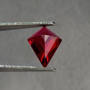 Three-quarter angle in tweezers of the same kite cut ruby, highlighting sparkle along the facet edges and tight geometry. The color is classic pigeon blood red, deeply saturated and vivid. This lab grown AAA grade ruby is precision cut in Thailand and offered loose for custom rings, necklaces, and fine one-of-a-kind settings.