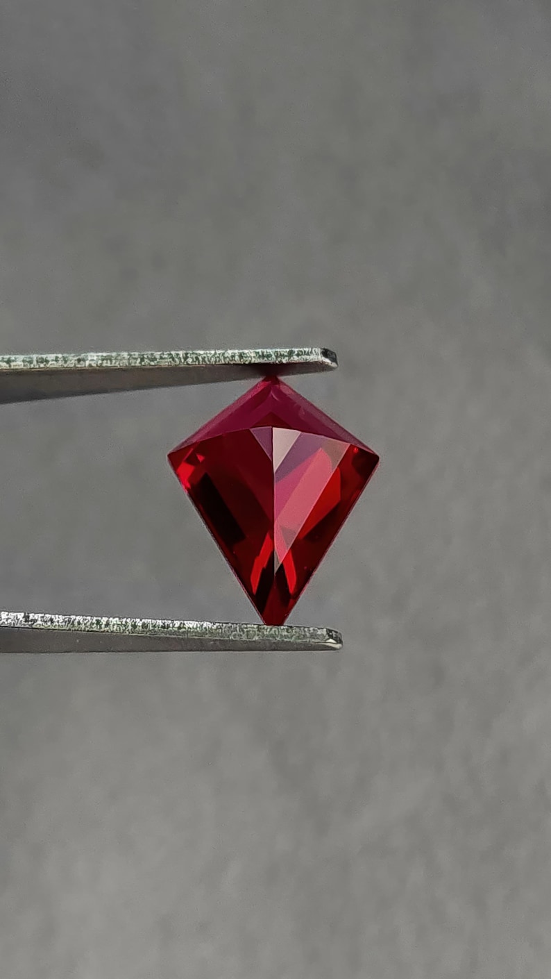 Three-quarter angle in tweezers of the same kite cut ruby, highlighting sparkle along the facet edges and tight geometry. The color is classic pigeon blood red, deeply saturated and vivid. This lab grown AAA grade ruby is precision cut in Thailand and offered loose for custom rings, necklaces, and fine one-of-a-kind settings.