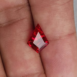 Hand shot of the same flawless ruby resting between fingers, tilted to show transparency and internal glow. The crisp kite silhouette and bright red saturation give a luxury look normally associated with very high-end natural ruby. Precision Thai faceting and excellent polish make this stone ready for bespoke fine jewelry.