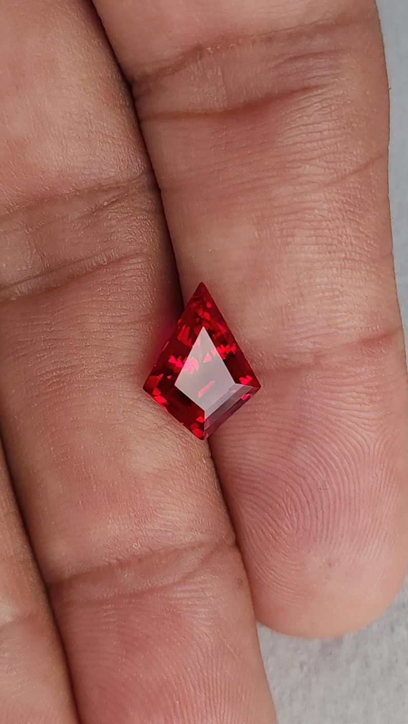 Hand shot of the same flawless ruby resting between fingers, tilted to show transparency and internal glow. The crisp kite silhouette and bright red saturation give a luxury look normally associated with very high-end natural ruby. Precision Thai faceting and excellent polish make this stone ready for bespoke fine jewelry.