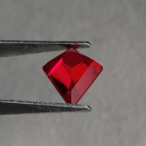 Side-angled view of the same lab grown pigeon blood red ruby in tweezers, showing the crisp crown and sharp outline of the kite / shield style cut. Bright internal reflections confirm flawless clarity and excellent light return. Superior Thai faceting and mirror polish highlight this AAA grade synthetic ruby, perfect for made-to-order fine jewelry or a standout solitaire.