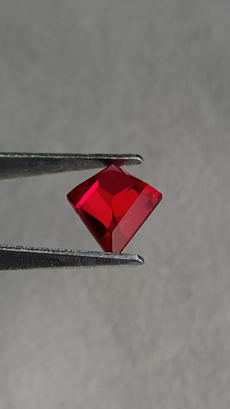 Side-angled view of the same lab grown pigeon blood red ruby in tweezers, showing the crisp crown and sharp outline of the kite / shield style cut. Bright internal reflections confirm flawless clarity and excellent light return. Superior Thai faceting and mirror polish highlight this AAA grade synthetic ruby, perfect for made-to-order fine jewelry or a standout solitaire.
