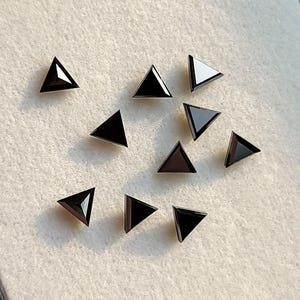 Puede incluir: Un conjunto de gemas triangulares negras, cada una con una superficie pulida y reflectante. Las gemas están dispuestas sobre una superficie clara y texturizada, mostrando su forma geométrica y color oscuro.
