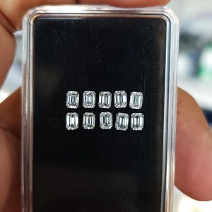 Hand holding a small clear plastic gem box with ten emerald cut diamonds on a black base, displaying the matched 3x2mm and 4x3mm calibrated stones.