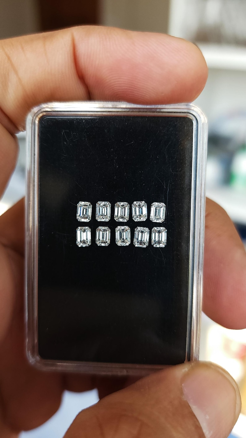 Hand holding a small clear plastic gem box with ten emerald cut diamonds on a black base, displaying the matched 3x2mm and 4x3mm calibrated stones.