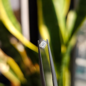 Natural emerald cut diamond placed on the pads of two fingers, emphasizing the compact size suitable as a side stone for engagement rings.