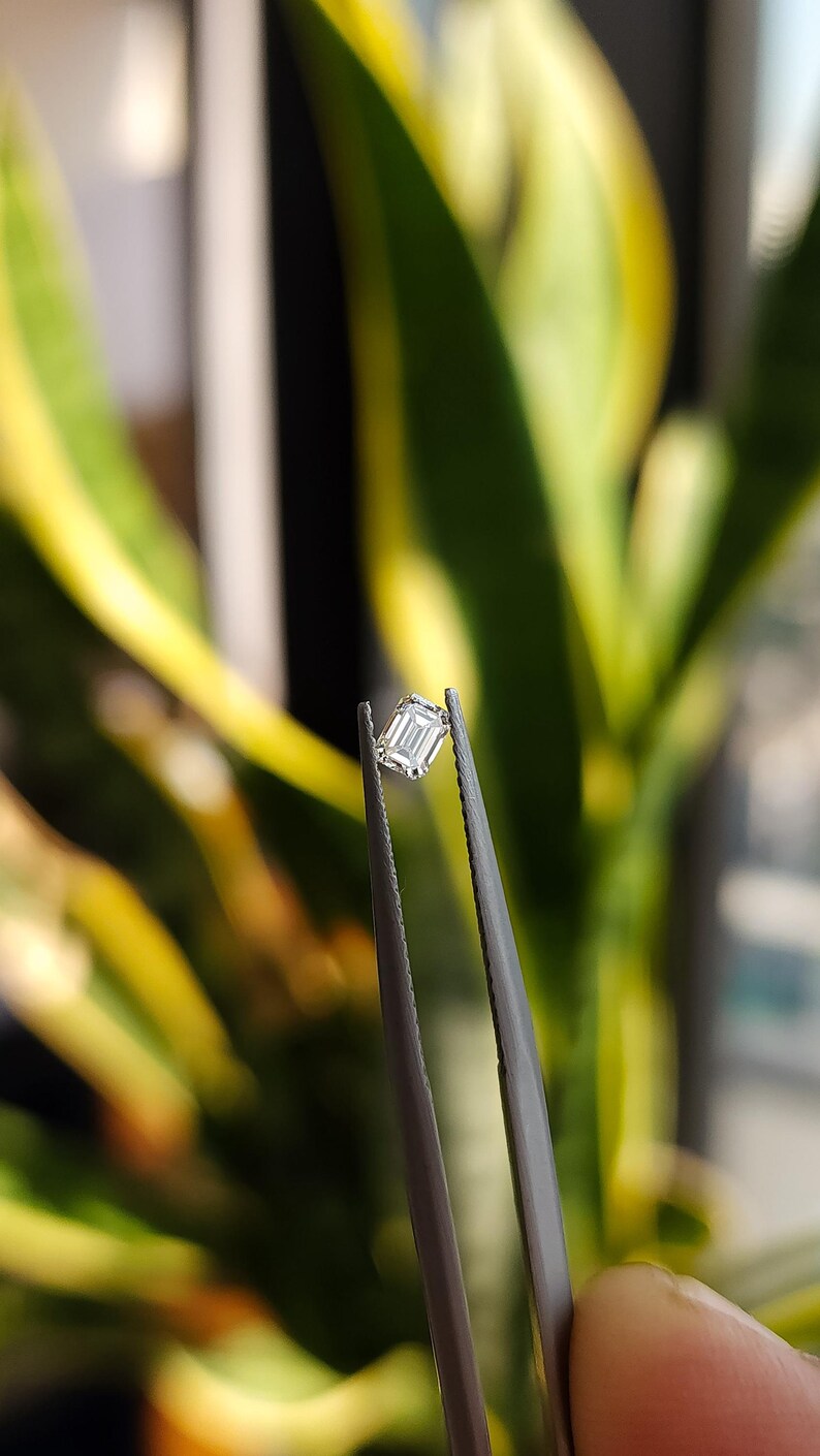 Natural emerald cut diamond placed on the pads of two fingers, emphasizing the compact size suitable as a side stone for engagement rings.