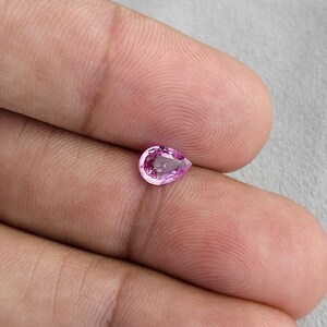 Fingertip scale photo of a small pear cut unheated natural sapphire, vivid orchid pink tone in natural light, clean outline and balanced teardrop proportions, Madagascar natural sapphire for a delicate engagement ring or pendant.