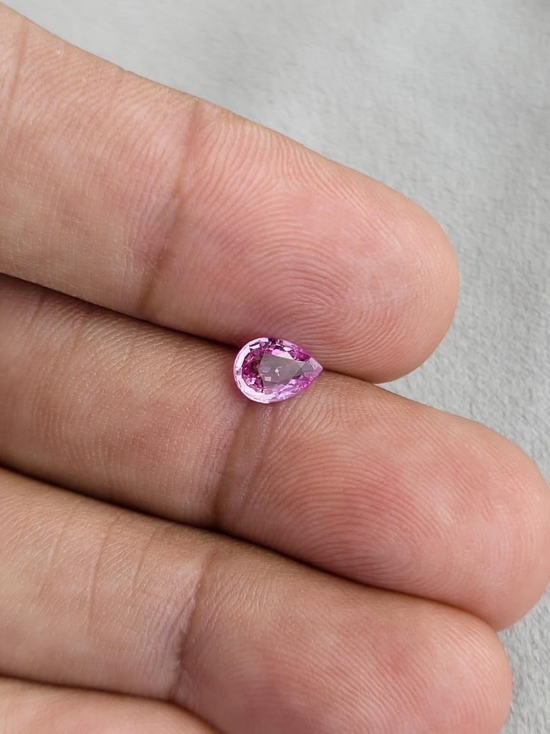 Fingertip scale photo of a small pear cut unheated natural sapphire, vivid orchid pink tone in natural light, clean outline and balanced teardrop proportions, Madagascar natural sapphire for a delicate engagement ring or pendant.