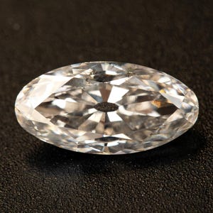 Può includere: Una gemma trasparente di forma ovale con molteplici sfaccettature che riflettono la luce. La gemma ha una forma leggermente allungata e poggia su una superficie scura. Il taglio crea un brillante gioco di luci e ombre.