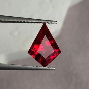 Face-up view of the kite cut ruby held in tweezers on a pale gray background. The stone flashes bright crimson highlights and deep pigeon blood cores, with flawless clarity and beautiful symmetry. A custom jeweler could easily bezel or prong set this lab created ruby as a luxury focal point in fine gold or platinum work.