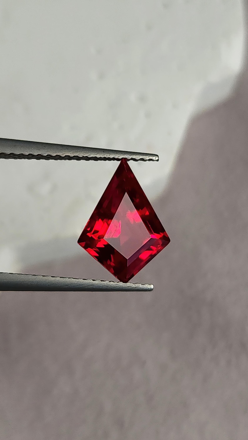 Face-up view of the kite cut ruby held in tweezers on a pale gray background. The stone flashes bright crimson highlights and deep pigeon blood cores, with flawless clarity and beautiful symmetry. A custom jeweler could easily bezel or prong set this lab created ruby as a luxury focal point in fine gold or platinum work.