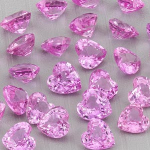 Puede incluir: Colección de gemas rosas facetadas en forma de corazón sobre una superficie gris. Las gemas, de varios tamaños, brillan intensamente. Ideales para la creación de joyas.