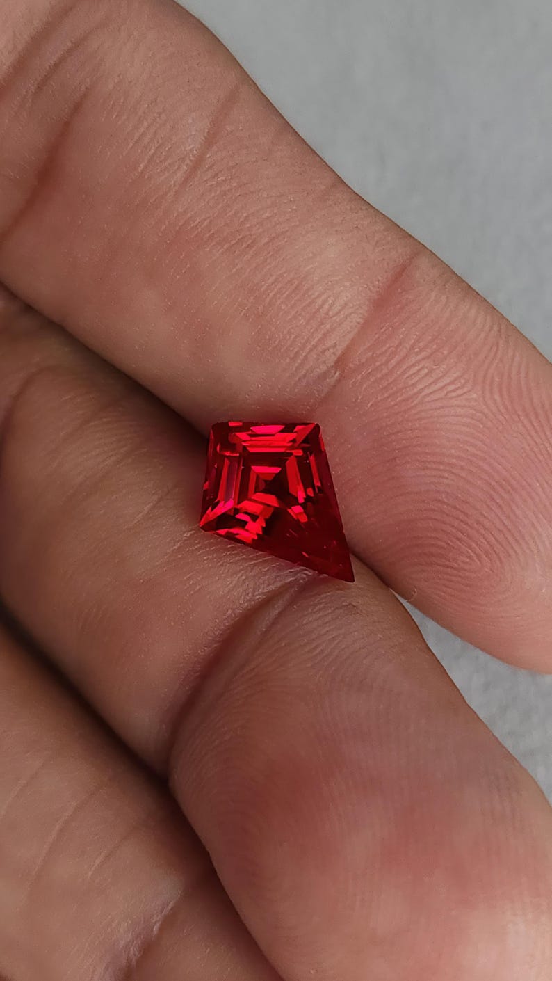 Close-up of the same kite cut ruby resting on skin at a slight angle, catching internal neon red fire. This photo highlights face-up brilliance and crisp facet junctions from expert Thai cutting. The stone is AAA grade, flawless clarity, and sold loose for custom jewelry orders, including unique engagement rings and luxury pendants.
