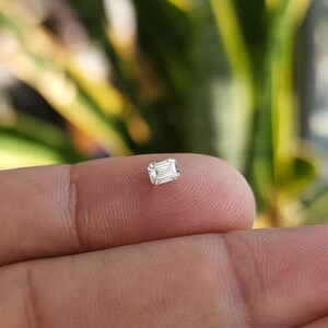 Single natural emerald cut diamond resting on a fingertip in outdoor light with blurred plants behind, showing bright white brilliance of the F G SI stone.