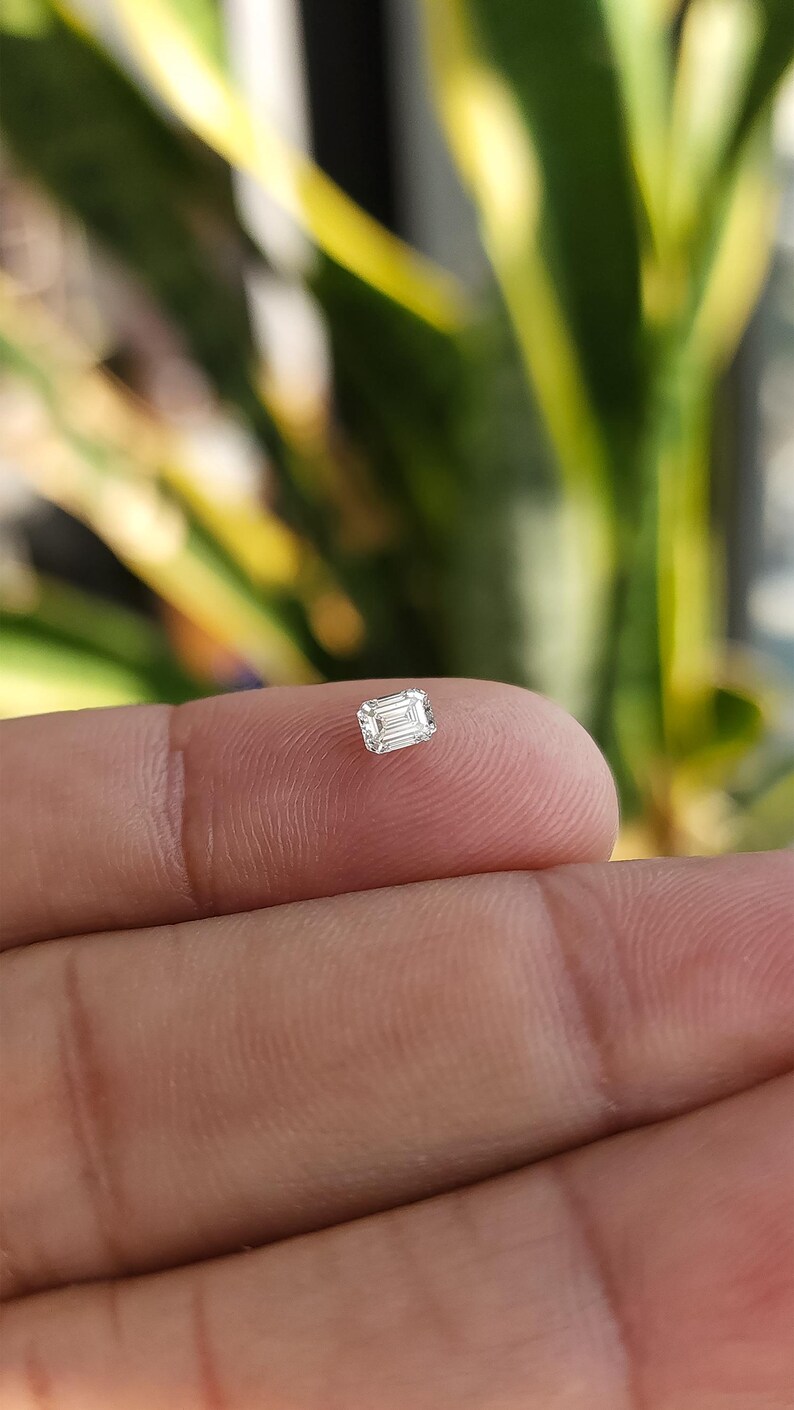 Single natural emerald cut diamond resting on a fingertip in outdoor light with blurred plants behind, showing bright white brilliance of the F G SI stone.