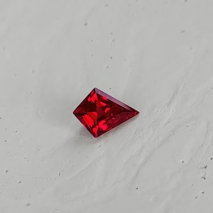 Angled shot of the same synthetic ruby on a pale surface, showing the crisp outline and polished facet edges. The vivid red color and internal brightness speak to high-grade lab grown material and expert faceting. A great option for clients who want bold ruby color, flawless clarity, and modern shape without paying natural ruby pricing.