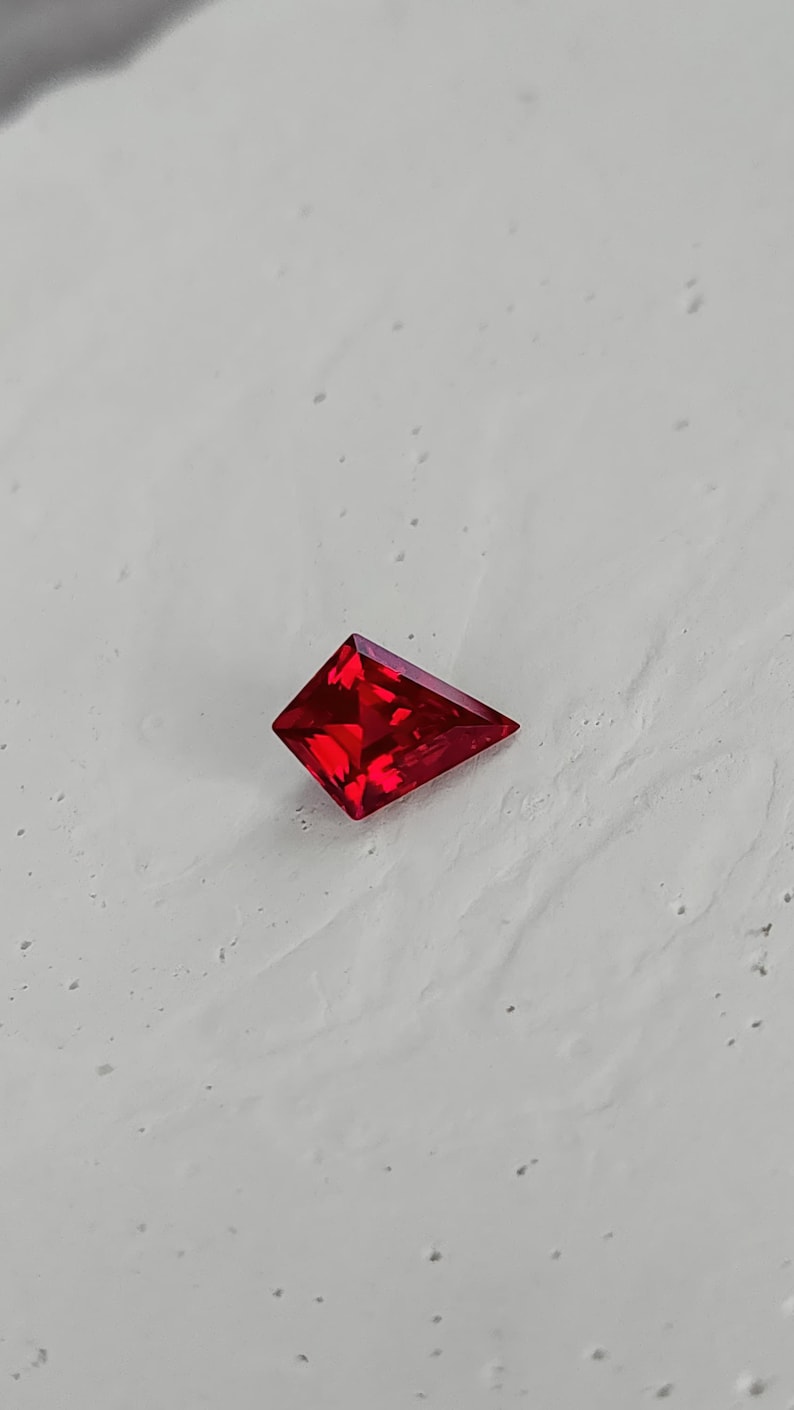 Angled shot of the same synthetic ruby on a pale surface, showing the crisp outline and polished facet edges. The vivid red color and internal brightness speak to high-grade lab grown material and expert faceting. A great option for clients who want bold ruby color, flawless clarity, and modern shape without paying natural ruby pricing.