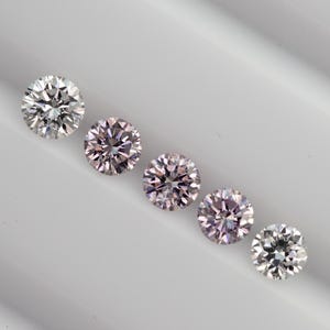 May include: Five round brilliant cut gemstones arranged in a diagonal line. The first and last stones are clear and colorless, while the three center stones exhibit a soft, pale pink hue. Each stone features multiple facets that reflect light.