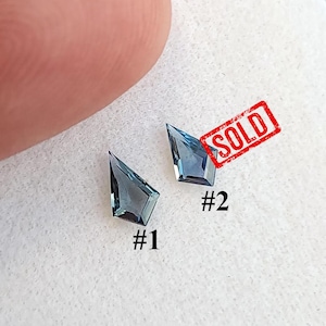 Puede incluir: Dos piedras preciosas azules en forma de cometa con superficies facetadas. Las piedras están etiquetadas como #1 y #2, con un sello rectangular rojo sobre la segunda piedra que dice SOLD. Las gemas exhiben un tono azul translúcido y bordes afilados.