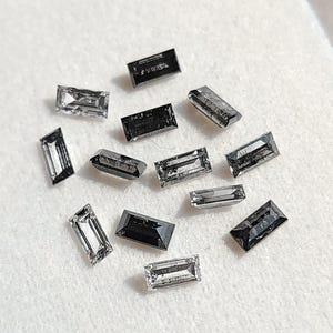 May include: A selection of rectangular-cut gemstones in shades of grey and black, arranged on a white surface. The gemstones have a reflective quality, creating a sparkling effect. The image showcases a collection of jewellery and gemstones.