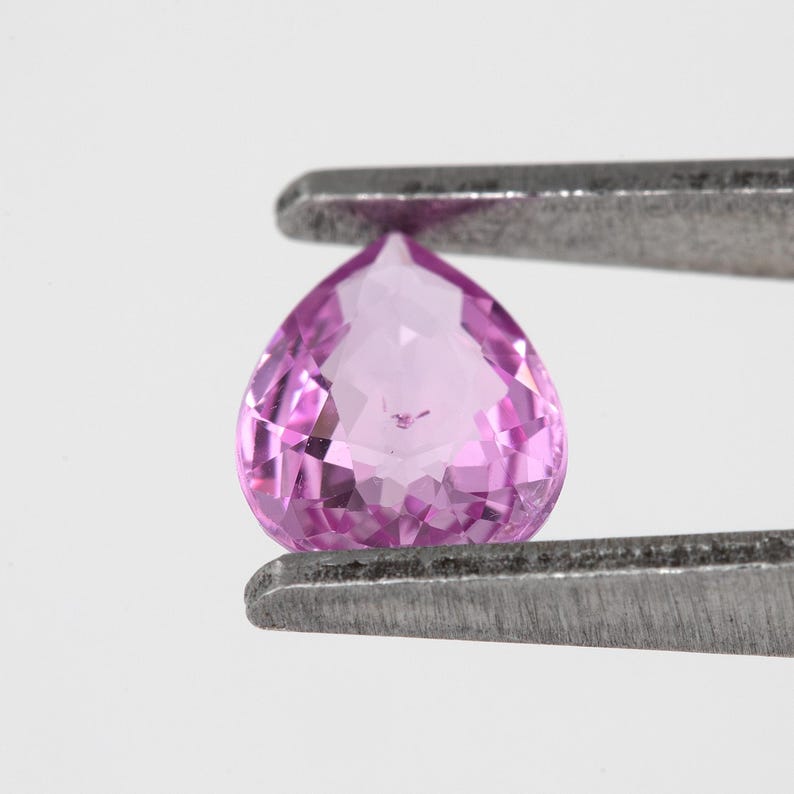 Side angle tweezers view of the pear cut unheated natural sapphire, vivid orchid pink body color through the stone, showing crown height and pavilion depth for low profile setting planning.