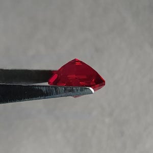 Profile shot of the precision-cut kite shape lab created ruby held in tweezers, showing pavilion depth and clean girdle. The stone glows intense pigeon blood red with zero visible inclusions, proof of flawless clarity. This custom-cut AAA grade ruby is calibrated for high-end rings, pendants, and bespoke settings where you want bold color without natural ruby pricing.