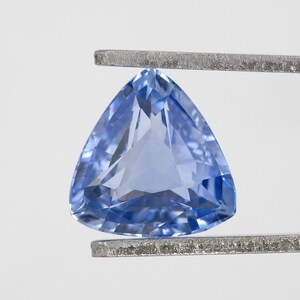 May include: A triangular, light blue gemstone held by a pair of silver tweezers. The gem has a faceted surface, catching the light and displaying various shades of blue. The shape is a perfect equilateral triangle.