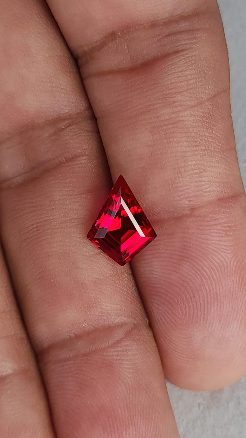 The kite cut lab grown ruby shown on fingertips for natural scale. The stone flashes intense pigeon blood red under soft light, with sharp step facets and flawless clarity. This loose AAA grade synthetic ruby is perfect for an engagement ring center stone, statement pendant, or matched pair earrings. Custom cutting and sizing services are available.