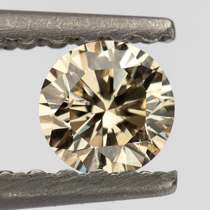 May include: A round, champagne-colored gemstone with a brilliant cut, featuring numerous triangular and kite-shaped facets that create a complex pattern of light reflection. The stone is held securely between the serrated jaws of metal tweezers.