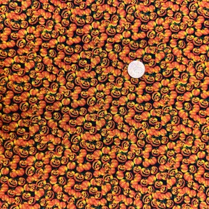 May include: Fabric with a repeating pattern of orange jack-o'-lantern faces on a black background. The pumpkins have carved faces and are in various sizes. A silver coin is in the center of the image.