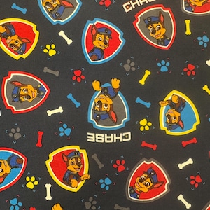 May include: Dark blue fabric featuring a repeating pattern of the character Chase from Paw Patrol. The design includes Chase in a shield, paw prints, and the word "CHASE" in white. The fabric also has bone shapes.