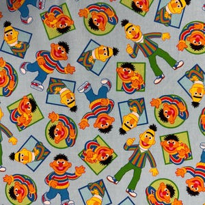 May include: Light blue fabric featuring a repeating pattern of the Sesame Street character Ernie. Ernie is depicted in various poses, wearing his signature striped shirt, green pants, and yellow hair. The design includes green circles and blue squares.