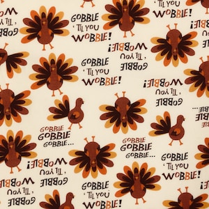 May include: Off-white fabric with a repeating pattern of cartoon turkeys in shades of brown and orange. The text "GOBBLE 'TIL YOU WOBBLE!" is interspersed throughout the design, creating a festive Thanksgiving theme.