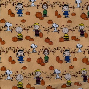 May include: A repeating pattern featuring cartoon characters, pumpkins, and autumn leaves on a light orange background. The characters include Snoopy, Charlie Brown, Lucy, and others. The pumpkins are orange, and the leaves are brown and yellow.