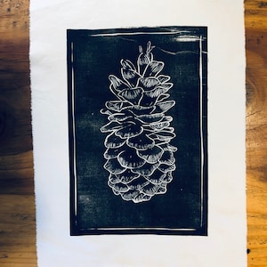 May include: A black and white linocut print of a pine cone. The print is framed with a black border and is on a white background.