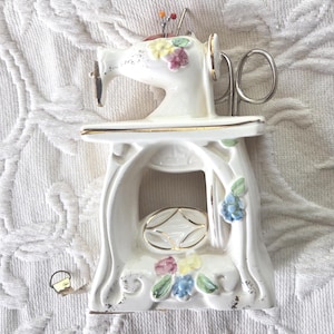 May include: A white ceramic sewing machine pin cushion with gold accents and floral details. The pin cushion has a decorative base with a gold oval design. Several pins are inserted into the top of the sewing machine.