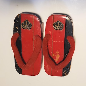 May include: Pair of traditional Japanese geta sandals. The wooden sandals feature a red and black color scheme with a decorative gold emblem. The thong straps are red. The geta are designed to elevate the wearer's feet.