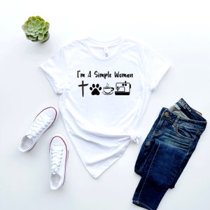 May include: White t-shirt with the text "I'm a simple woman" and four icons: a cross, a paw print, a cup of coffee, and a sewing machine.