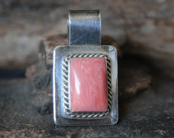 May include: A silver pendant with a pink and white stone set in a silver frame. The pendant is on a rough, brown surface.