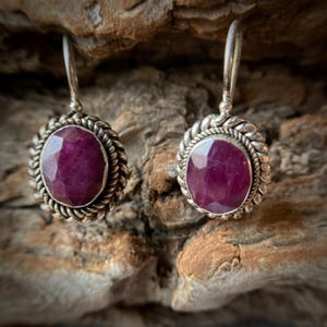 May include: A pair of silver earrings with a dark pink oval gemstone set in a twisted silver frame.
