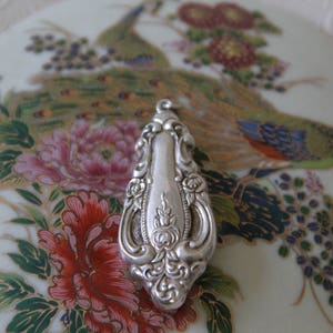May include: A silver pendant in the shape of a teardrop with intricate floral designs. The pendant is on a white background with a floral pattern.