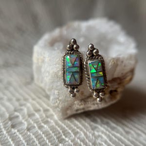 May include: A pair of rectangular stud earrings featuring inlaid iridescent opal segments in a geometric pattern. The silver settings have a twisted rope border and small decorative spheres at the top and bottom of each earring, showcasing a metallic finish.